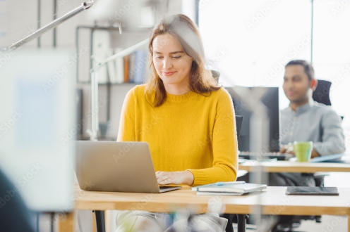 Nuori nainen keltaisessa puserossa työskentelee etualalla läppärillä ja mies taka-alalla olevassa pöydässä.